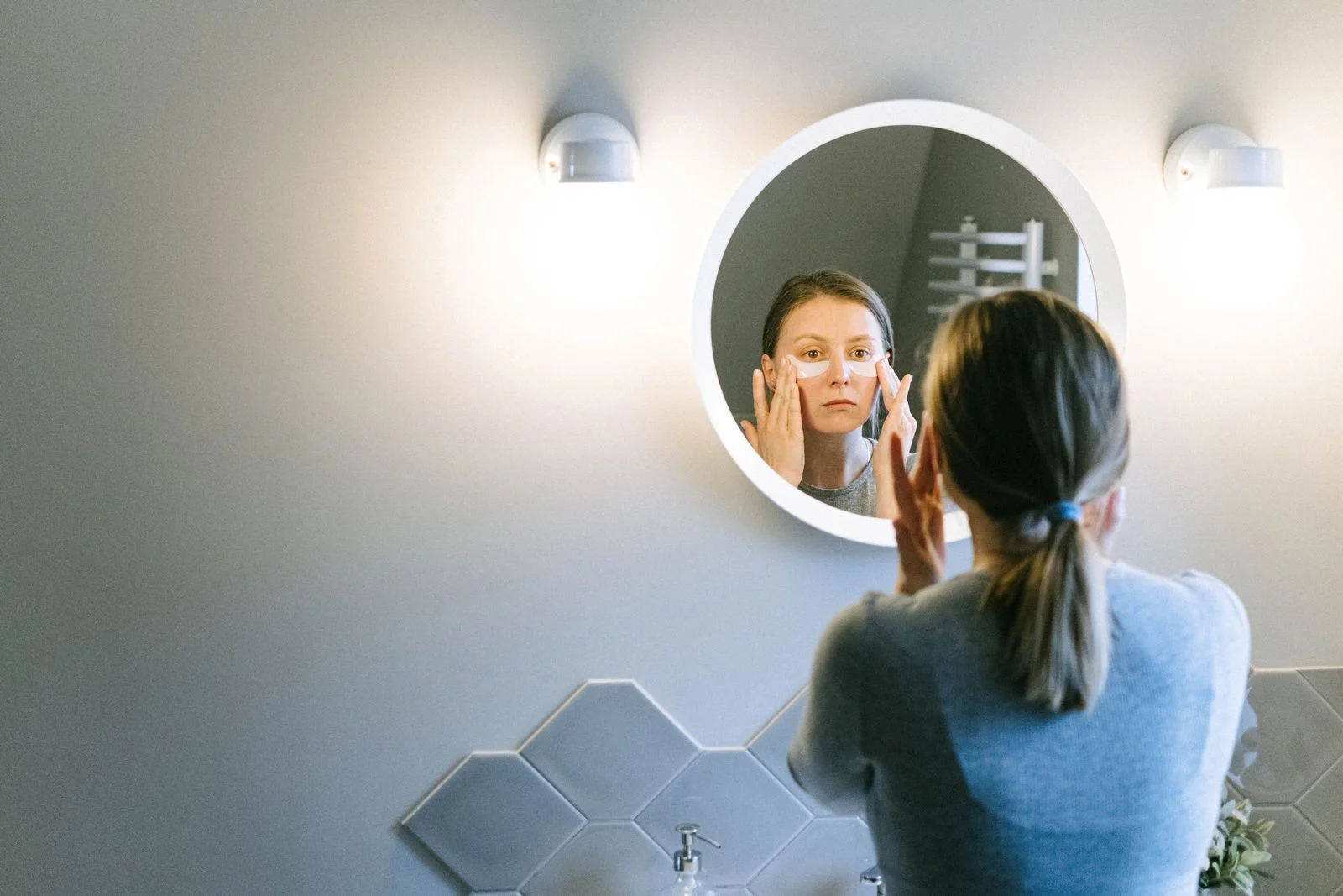 Femme qui se masse le visage devant un miroir rond éclairé de salle de bain.
