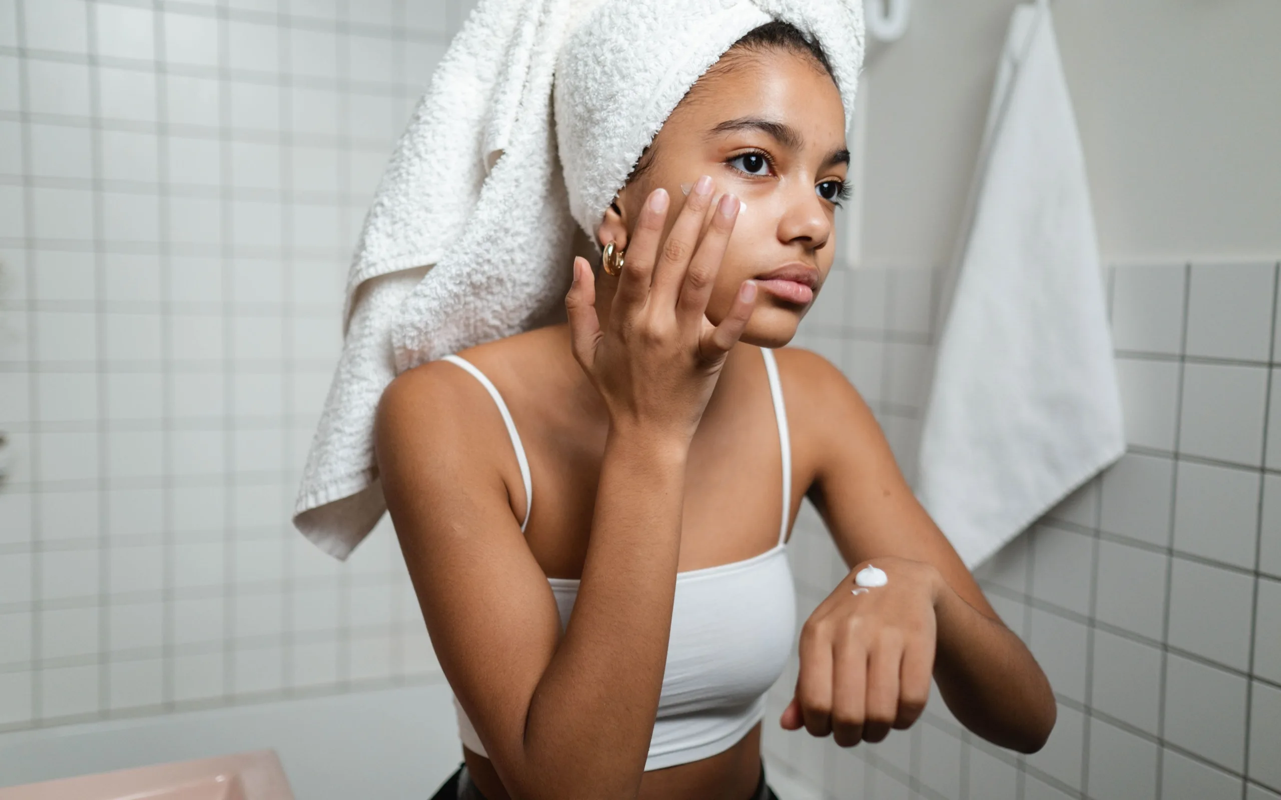Jeune femme, serviette sur les cheveux, qui applique sa crème hydratante devant le miroir de la salle de bain.